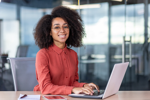 Portrait of successful businesswoman at workplace inside office, woman smiles and looks at camera, uses laptop at work, female programmer in red shirt with curly hair, codes new software.