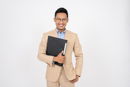 A smart and happy Asian businessman in a formal suit with a document binder stands on an isolated white background. businesspeople, corporate job, business entrepreneur