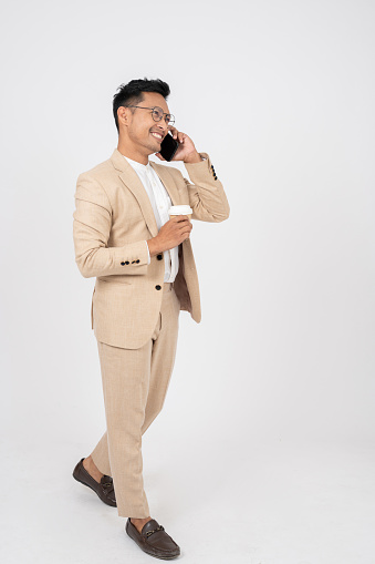 A smart and positive Asian businessman in a formal suit is talking on the phone and walking with a coffee cup in his hand, on an isolated white studio background.
