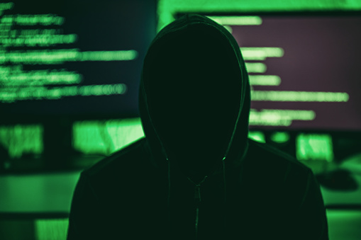 Anonymous internet hacker in front of computer monitors. Obscured face.