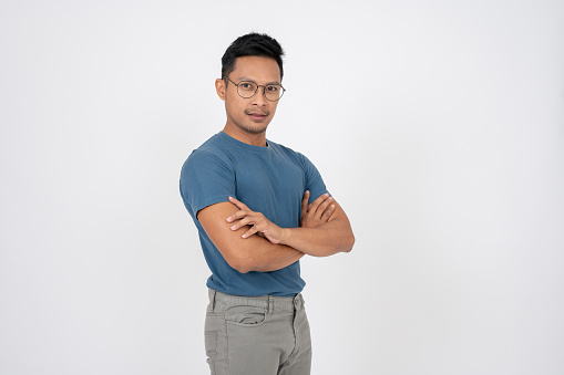 A strict, serious Asian man in casual clothes is standing on an isolated white studio background with his arms crossed.