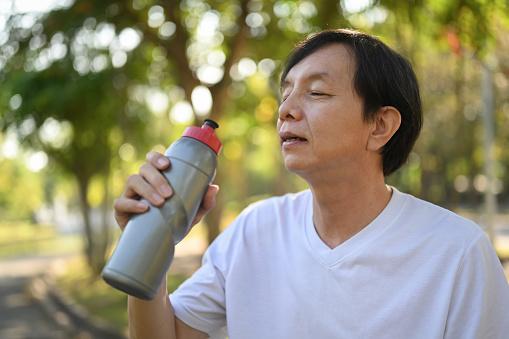 Senior man athlete drinking water from a bottle, resting after jogging outdoors on warm sunny day.
