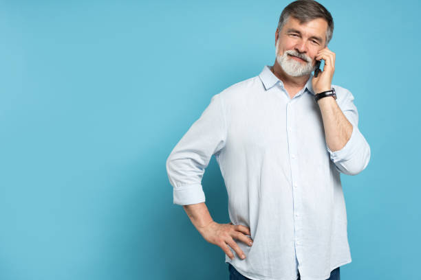 istock Happy mature bearded man standing over blue wall background isolated. Looking aside while talking by phone 1866802592
