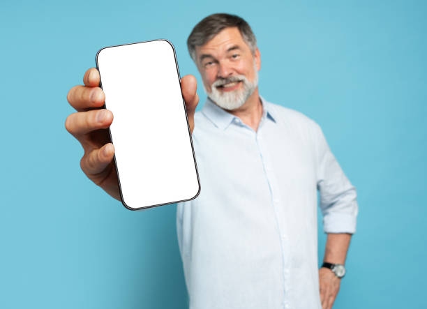 istock Handsome Excited Mature Man Showing Pointing At Empty Smartphone Screen Posing Over Light Blue Studio Background, Smiling To Camera. Check This Out, Cellphone Display Mock Up 1866802595