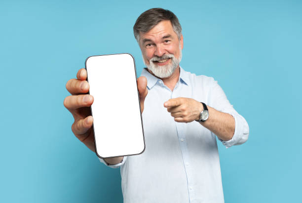 istock Handsome Excited Mature Man Showing Pointing At Empty Smartphone Screen Posing Over Light Blue Studio Background, Smiling To Camera. Check This Out, Cellphone Display Mock Up 1866802596