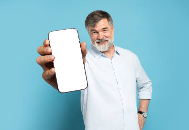 istock Handsome Excited Mature Man Showing Pointing At Empty Smartphone Screen Posing Over Light Blue Studio Background, Smiling To Camera. Check This Out, Cellphone Display Mock Up 1866802605