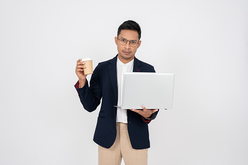 A stressed, serious Asian businessman in a formal suit is standing on an isolated white background with his laptop computer and a coffee cup in his hand.