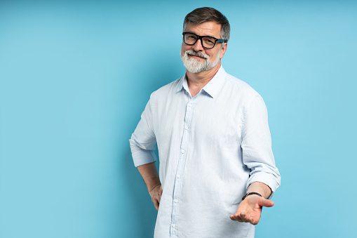 Bearded middle-aged man wearing glasses posing over blue studio background with copy space looking at the camera with a friendly smile