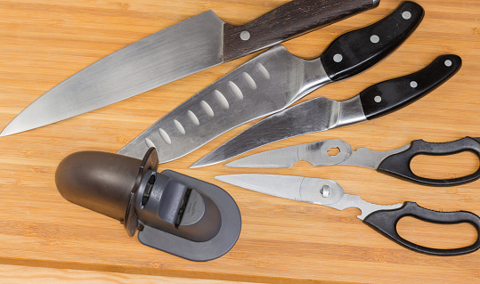 Used manual kitchen combined sharpener of broach type on a blurred background of the different old kitchen knives and scissors on the wooden cutting board, top view