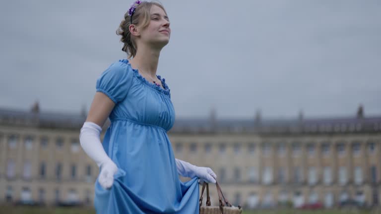 Young woman wearing a regency era dress is walking in a public park in front of Royal Crescent
