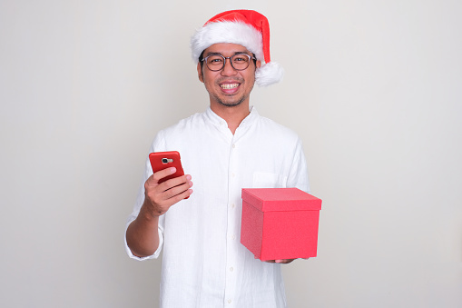 Asian man wearing christmas hat smiling with hand holding phone and gift box