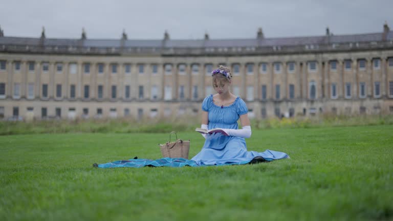 Young woman wearing a regency era dress is reading a book in a public park