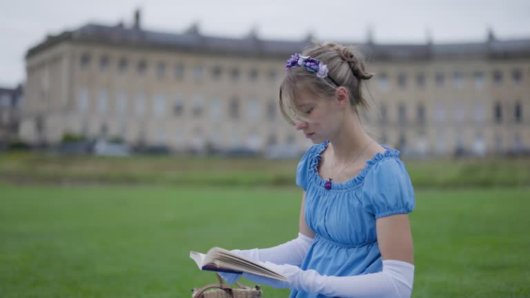 Young woman wearing a regency era dress is reading a book in a public park