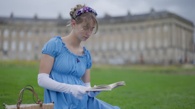 Young woman wearing a regency era dress is reading a book in a public park