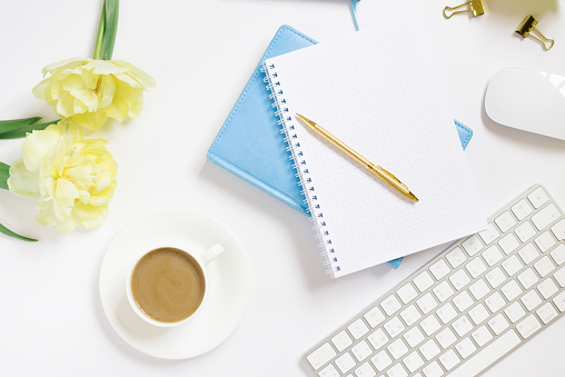 Notepad with a gold pen, yellow tulip flowers, a cup of coffee, a keyboard and a cup of coffee on a white background. Stylish workplace of a blogger or freelancer