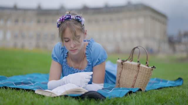 Young woman wearing a regency era dress is reading a book in a public park