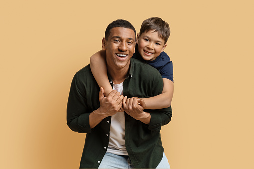 Happy black man have fun posing with cute child preteen boy. Daddy and son embracing and smiling isolated on beige background studio portrait. Fathers Day love family parenthood childhood concept