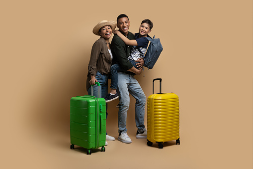 Family trip concept. Excited african american parents travelling with their preteen son, posing together with luggage suitcases and backpacks over beige background in studio, empty space