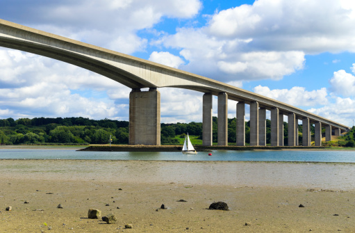 A small dinghy sailing upstream below the Orwell Bridge, which crosses the River Orwell at Wherstead, near Ipswich in Suffolk, England. 