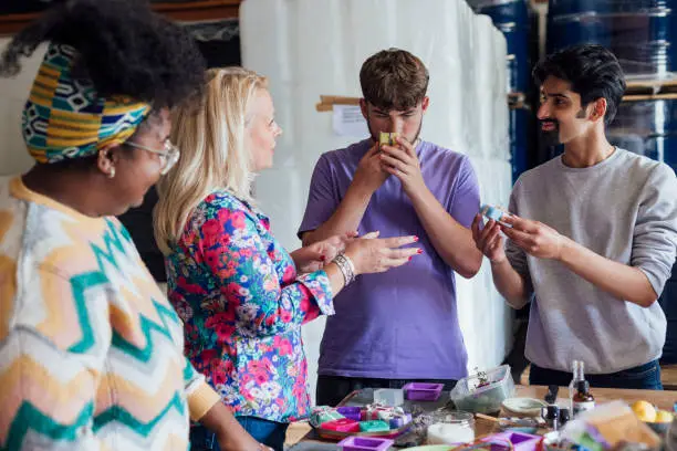 A shot of a group of people taking part in a soap making workshop. The workshop is being run by an independent business owner, and is taking place within an ink factory in Hexham, North East England. The group of people are standing around a table which is laid with materials and equipment, whilst they smell different herbs.