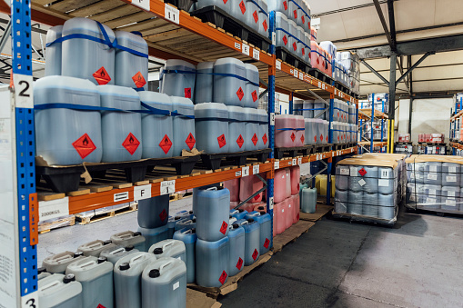 A shot of shelving units filled with plastic containers of ink in an ink factory in Hexham, North East England. The space is an industrial warehouse, and there are more containers of ink wrapped in plastic on pallets beside the shelving units.
