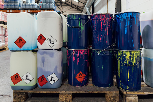 A shot of a wooden pallet stacked with plastic containers and metal drums of different coloured ink in an ink factory in Hexham, North East England. They are in an industrial warehouse, there are shelves full of more containers in partial view behind.