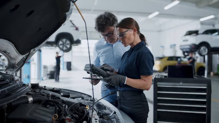 Female mechanic running diagnostic test on car engine in repair shop. Manager Checks Data on a Tablet and Explains an Engine Breakdown to a Handsome Professional Mechanic. Car Service Employees Inspect a Car with Internal Combustion Engine.