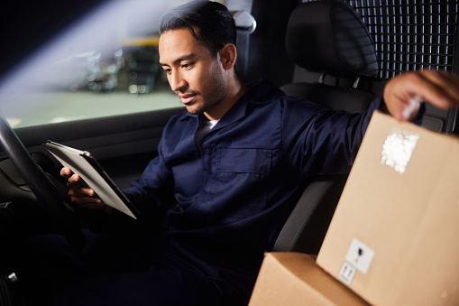 Young male delivery person using a mobile app on a digital tablet while sitting in his van before leaving a shipping and distribution warehouse