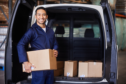 Portrait of a smiling young male delivery person loading his van with boxes before leaving a shipping and distribution warehouse