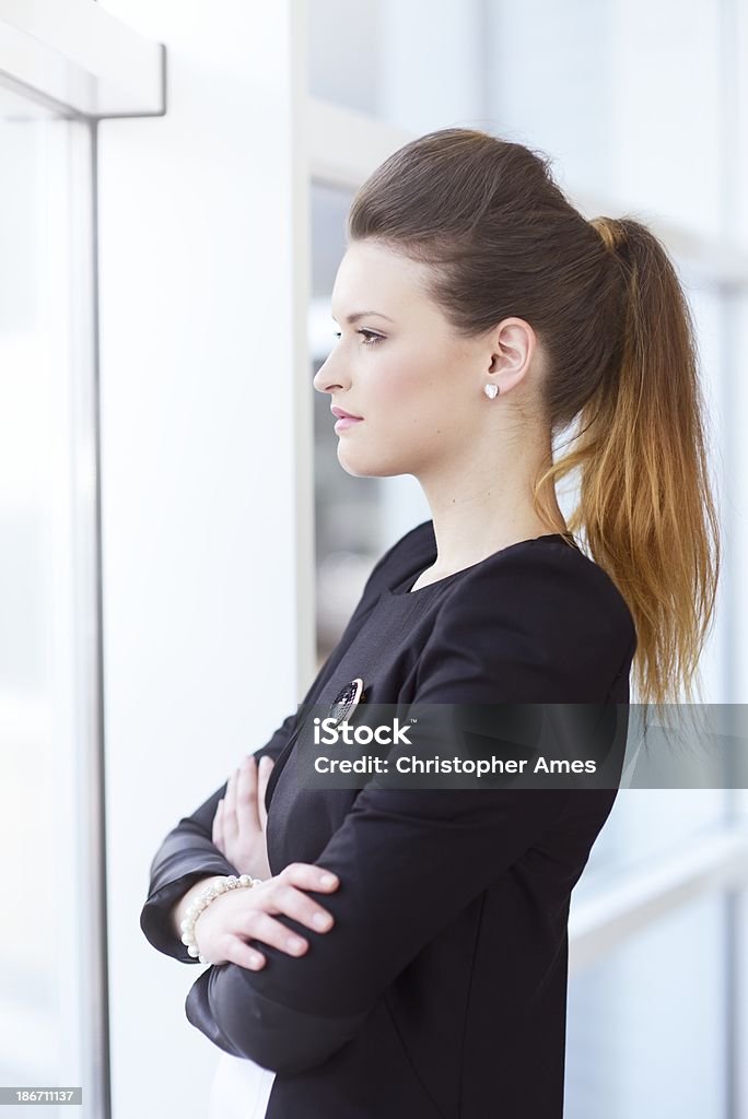 Confident Female Office Worker Looking Through Window A successful young businesswoman with crossed arms stands at the window of a modern office building and contemplates. 20-24 Years Stock Photo