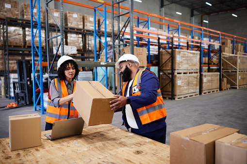 Two workers reading a bar code with a scanner and laptop while preparing packages for delivery on a table in a large shipping and distribution warehouse