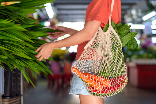 A cropped image captures an Asian woman using an eco-friendly tote bag while shopping for fresh vegetables and fruits at a local market stall, embodying the zero-waste concept