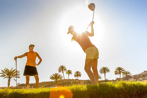 A young male golfer is silhouetted as he swings against a background of blue sky and trees. Copy space on clear blue sky and lens flare from rising sun..