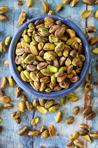 Stock photo showing elevated view of shelled pistachio nuts piled high in a blue dish against a blue woodgrain background. Raw pistachios are considered to be a very healthy snack food and are high in vitamin B6, potassium, antioxidants and protein, boasting a list of health benefits and may aid weight loss.
