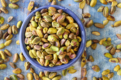 Stock photo showing elevated view of shelled pistachio nuts piled high in a blue dish against a blue woodgrain background. Raw pistachios are considered to be a very healthy snack food and are high in vitamin B6, potassium, antioxidants and protein, boasting a list of health benefits and may aid weight loss.