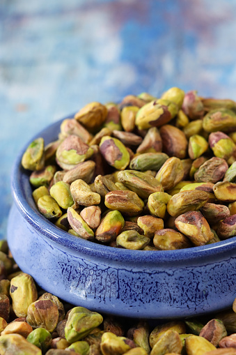 Stock photo showing close-up, elevated view of shelled pistachio nuts piled high in a blue dish against a blue woodgrain background. Raw pistachios are considered to be a very healthy snack food and are high in vitamin B6, potassium, antioxidants and protein, boasting a list of health benefits and may aid weight loss.