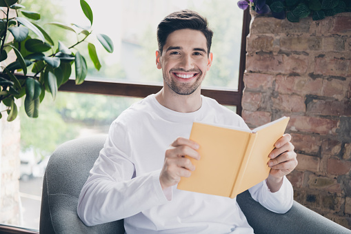 Photo of funky cheerful young guy wear white shirt reading interesting novel indoors apartment room.