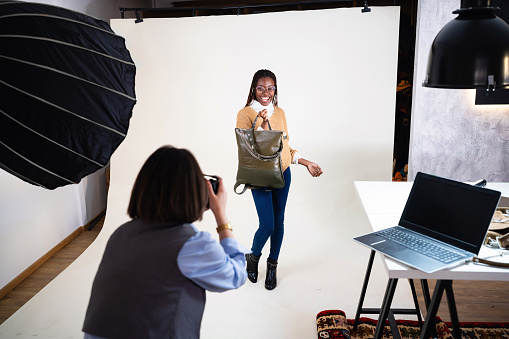 At the studio, young Caucasian female photographer, taking photos of the female fashion model Black ethnicity, while posing with an leather bag