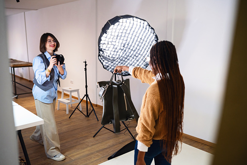 At the studio, young Caucasian female photographer, taking photos of the female fashion model Black ethnicity, while posing with an leather bag