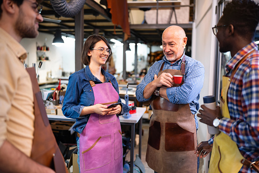 Senior Caucasian male tailor and group of diverse female and male trainees, talking and drinking coffee  at the leather shop