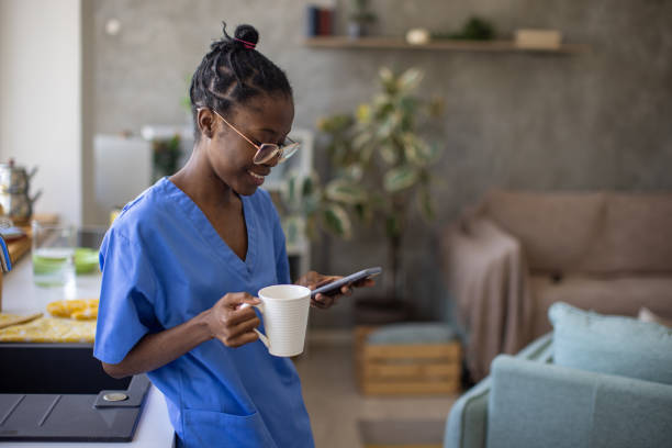Young woman wearing medical scrubs using her phone and drinking coffee at home