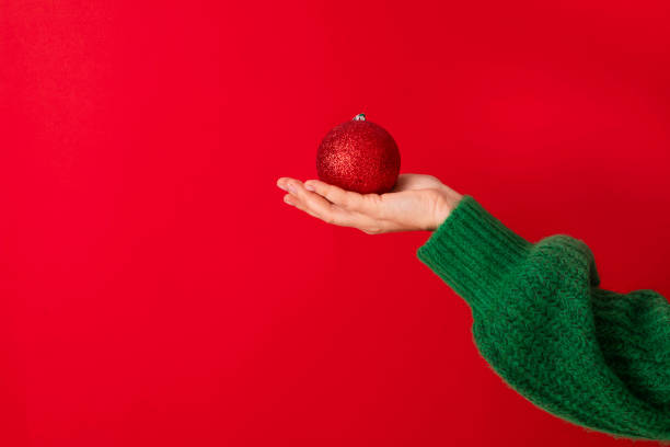 donne in un maglione verde che tiene in mano il simbolo del nuovo anno: una palla di natale rossa. - christmas fashion model human arm beautiful foto e immagini stock