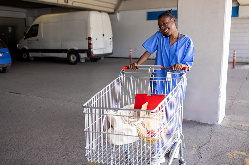 A young Black woman wearing medical scrubs loading groceries into a car