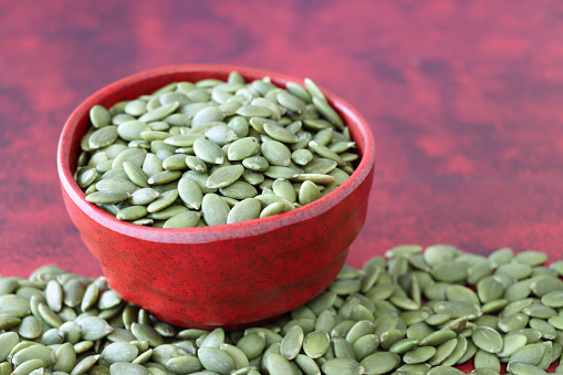 Stock photo showing close-up, elevated view of some pumpkin seeds, also known as pepita, piled high in a red dish against a mottled red background. Raw pumpkin seeds are considered to be a very healthy snack food and are high in vitamin K, manganese, antioxidants and protein, boasting a list of health benefits and may aid bladder health.