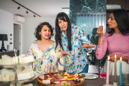 Young adult women of diverse backgrounds gather around a baby shower table adorned with a sumptuous selection of cheeses, fruits, and baked goods.