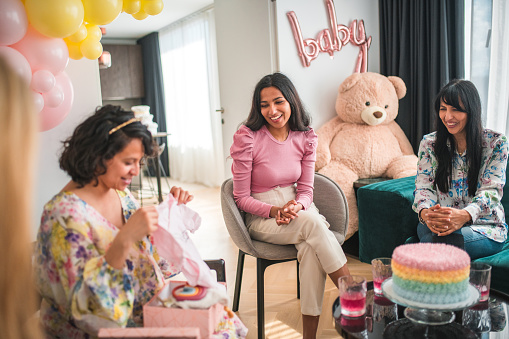 Generosity flows as a mid-adult woman unwraps a baby shower gift, encircled by the supportive presence of her diverse friends.