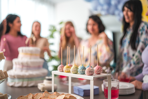 Colorful cake pops take center stage with a backdrop of laughing diverse women, including Hispanic and Indian, at a baby shower.