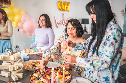 Mid adult women of diverse backgrounds gather around a baby shower table adorned with a sumptuous selection of cheeses, fruits, and baked goods.