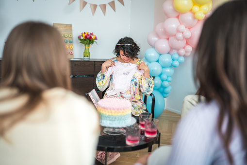 An expectant mother in a bright dress laughs with a diverse group of friends at her baby shower, surrounded by cheerful, pastel-colored decorations.