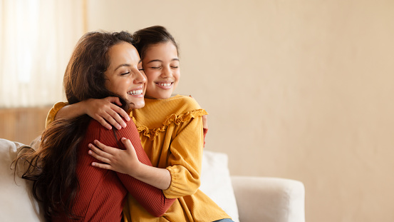 Cheerful cute hispanic preteen daughter girl hugging millennial mom, expressing their love and enjoying spare time in modern living room interior. Panorama, free space. Mother's support and care